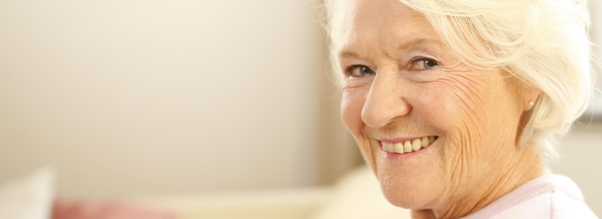 Lächelnde ältere Frau mit blonden Haaren, blickt in die Kamera, sits auf einem Sofa in heller Wohnumgebung.