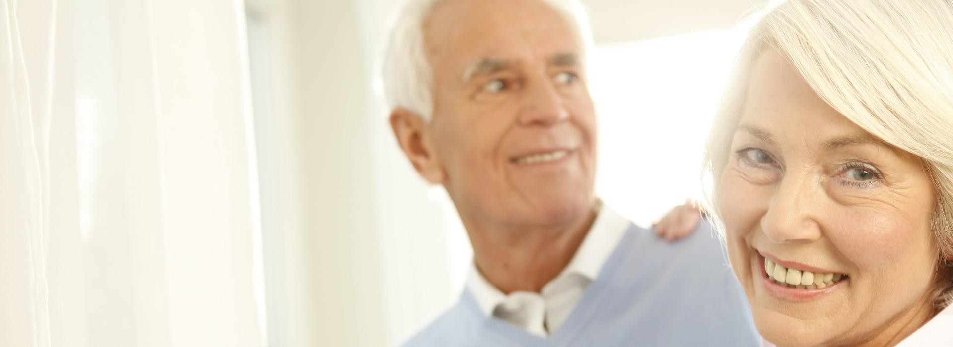 Ein älteres Paar steht in einem hellen Raum, lächelt und schaut sich an, Kleidung in sanften Farben.