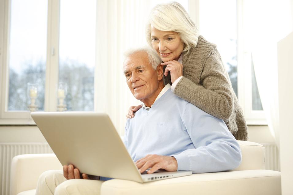 Älteres Paar sitzt auf einem Sofa und schaut gemeinsam auf einen Laptop, mit einem hellen Raum im Hintergrund.
