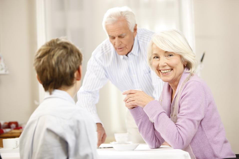 Lächelnde ältere Frau mit Kaffee, während ein älterer Mann im Hintergrund für ein Kind am Tisch sorgt.