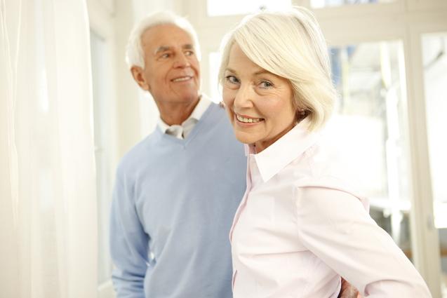 Ein lächelndes älteres Paar blickt sich an, im Hintergrund große Fenster und helle Vorhänge.
