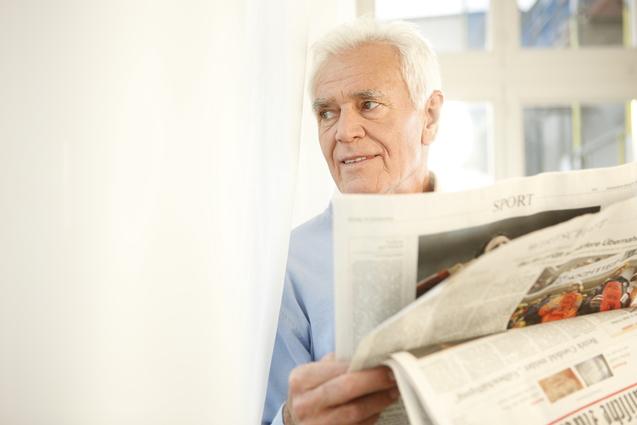 Älterer Mann mit weißem Haar liest eine Zeitung vor einem hellen Fenster.